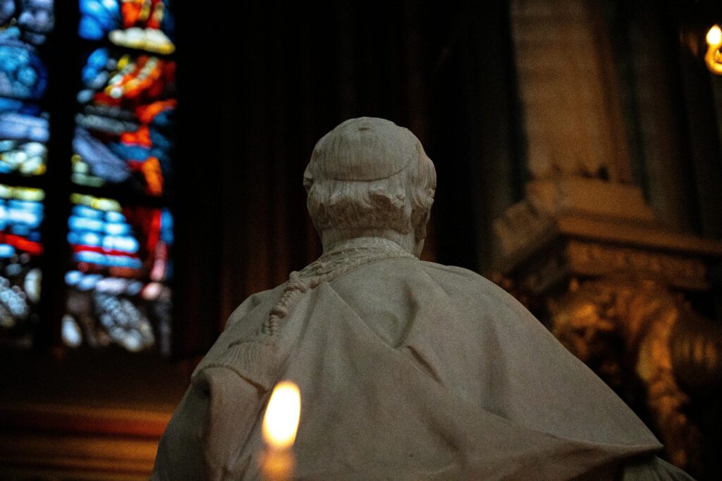 A serene religious statue silhouetted against vibrant stained glass in a dimly lit church interior.